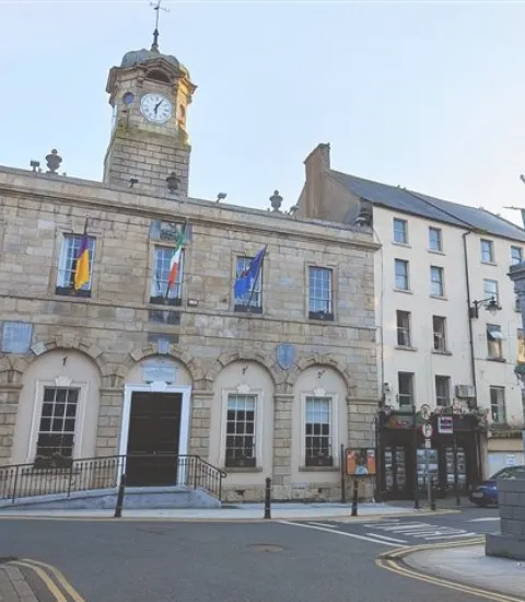 A historic stone building with arched windows and a clock tower, displaying four flags. Adjacent is a taller building with multiple windows and a pub at street level. The sky is clear, and a few bollards line the foreground.