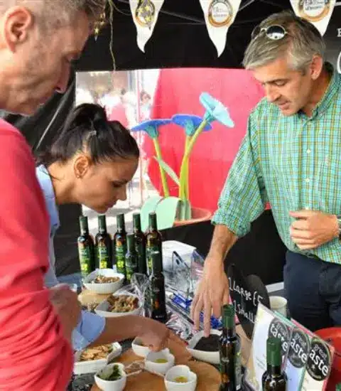 Three people at a market stall sample various foods. A woman dips bread into a dish while a man in a plaid shirt explains the products. Bottles lined up on the table suggest oils or vinegars. Colorful decorations are visible in the background.