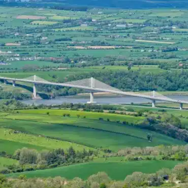 Aerial view of a modern cable-stayed bridge spanning a wide river in a lush, green rural landscape. Rolling hills and fields surround the river, and a distant horizon shows more greenery and scattered buildings.