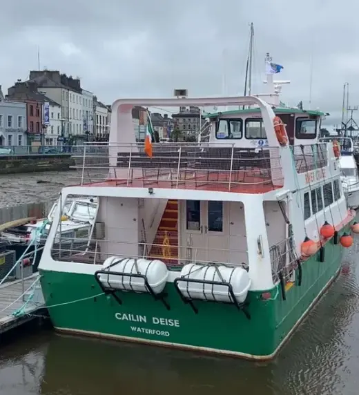 A green and white ferry named "Cailin Deise" is docked at a waterfront. There are buildings along the quay, and lifebuoys and a ladder are visible on the boat's deck. A small flag flutters on top, with a cloudy sky in the background.