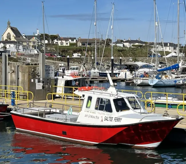 A small red and white boat named "Saltee Ferry" is docked at a marina. Several sailboats and a few buildings are visible in the background under a clear blue sky.