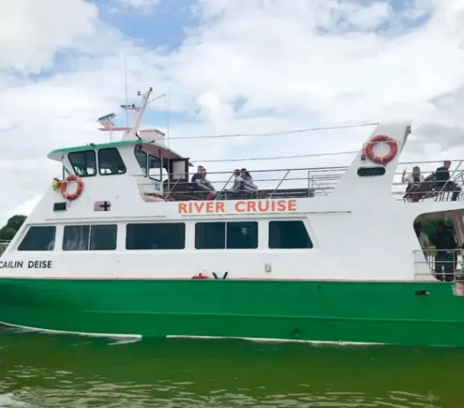 White and green river cruise boat labeled "Cailin Deise" with "River Cruise" on the side, floating on water. Several passengers can be seen on deck, with cloudy skies in the background.