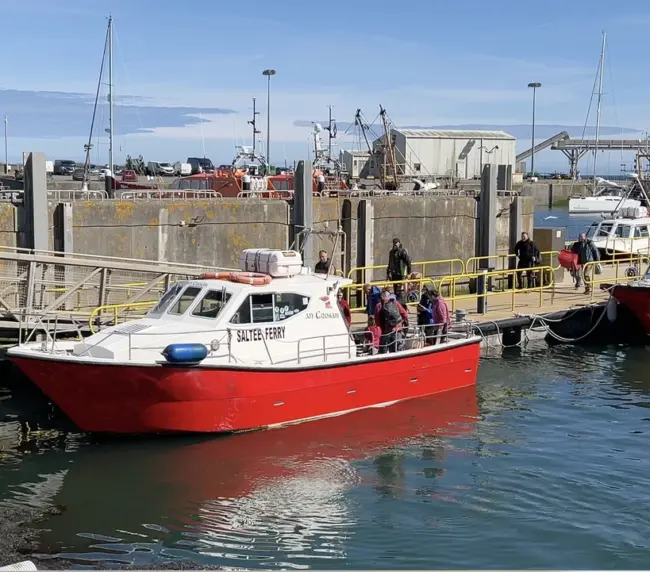 A red and white passenger boat docked at a harbor with people boarding. Other boats and industrial buildings are visible in the background under a clear blue sky.