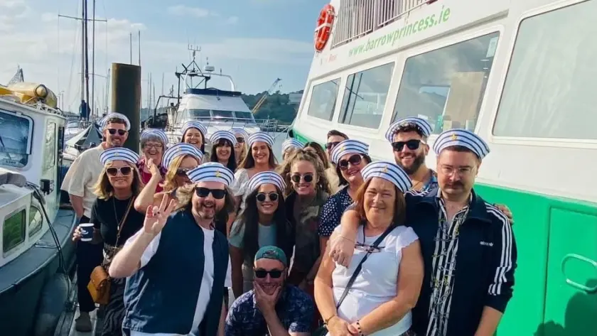 A group of people wearing sailor hats pose happily together on a dock beside a green and white boat. The sky is bright and partly cloudy, with other boats visible in the background.