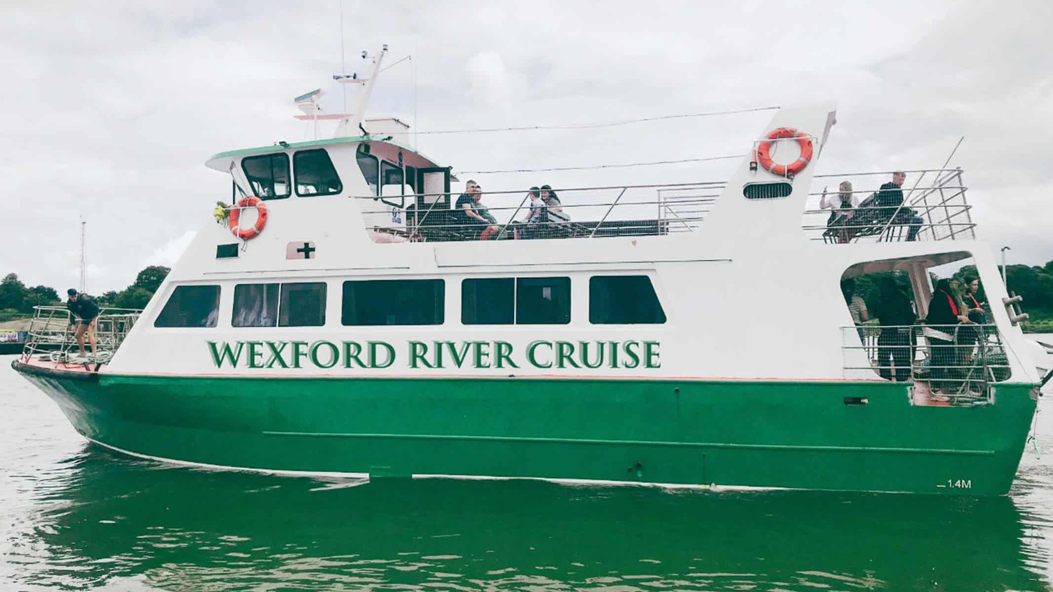 A green and white boat labeled "Wexford River Cruise" on the water. Several passengers are on the deck, some taking photos. Red lifebuoys are mounted on the sides. The background shows a cloudy sky and a distant treeline.