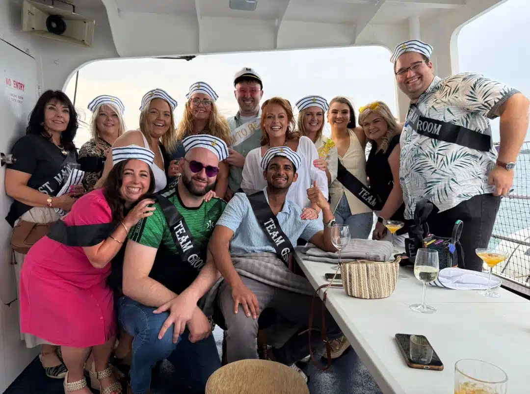 A group of people wearing striped sailor hats and black sashes smile and pose on a boat. Some sit, others stand. Food and drinks are on the table, and water is visible through the windows behind them.