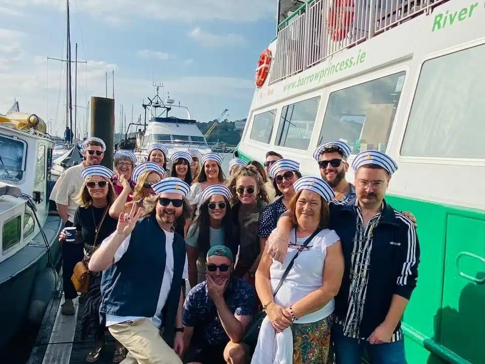 A group of people wearing matching sailor hats smile and pose together on a sunny quay next to a green and white boat. Some hold drinks and everyone looks cheerful, with water and more boats in the background.