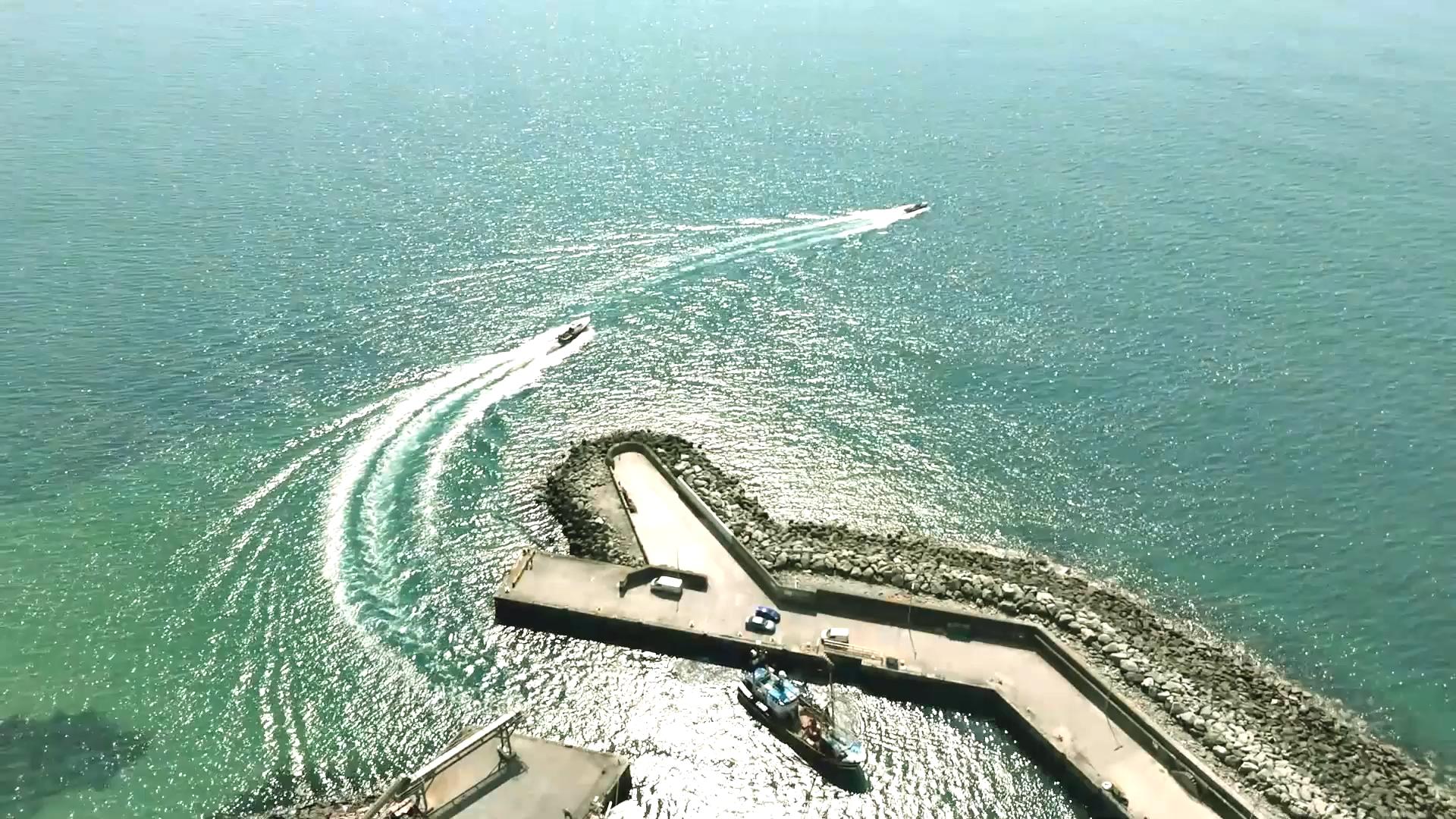 Aerial view of a pier extending into a sparkling turquoise sea. Several boats leave white trails in the water as they move near the breakwater. The sky is clear, and sunlight glistens on the ocean surface.